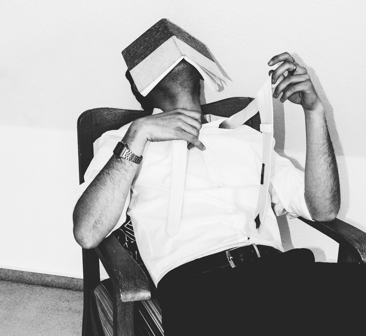 Black and white photograph of a man dressed in a white shirt and suspenders, reclining in a wooden chair. A thick book is resting open on his face, obscuring his identity. His tie is loosely draped over his shoulder, and one hand is raised in a relaxed gesture. The scene conveys exhaustion or frustration, possibly after studying or working late.